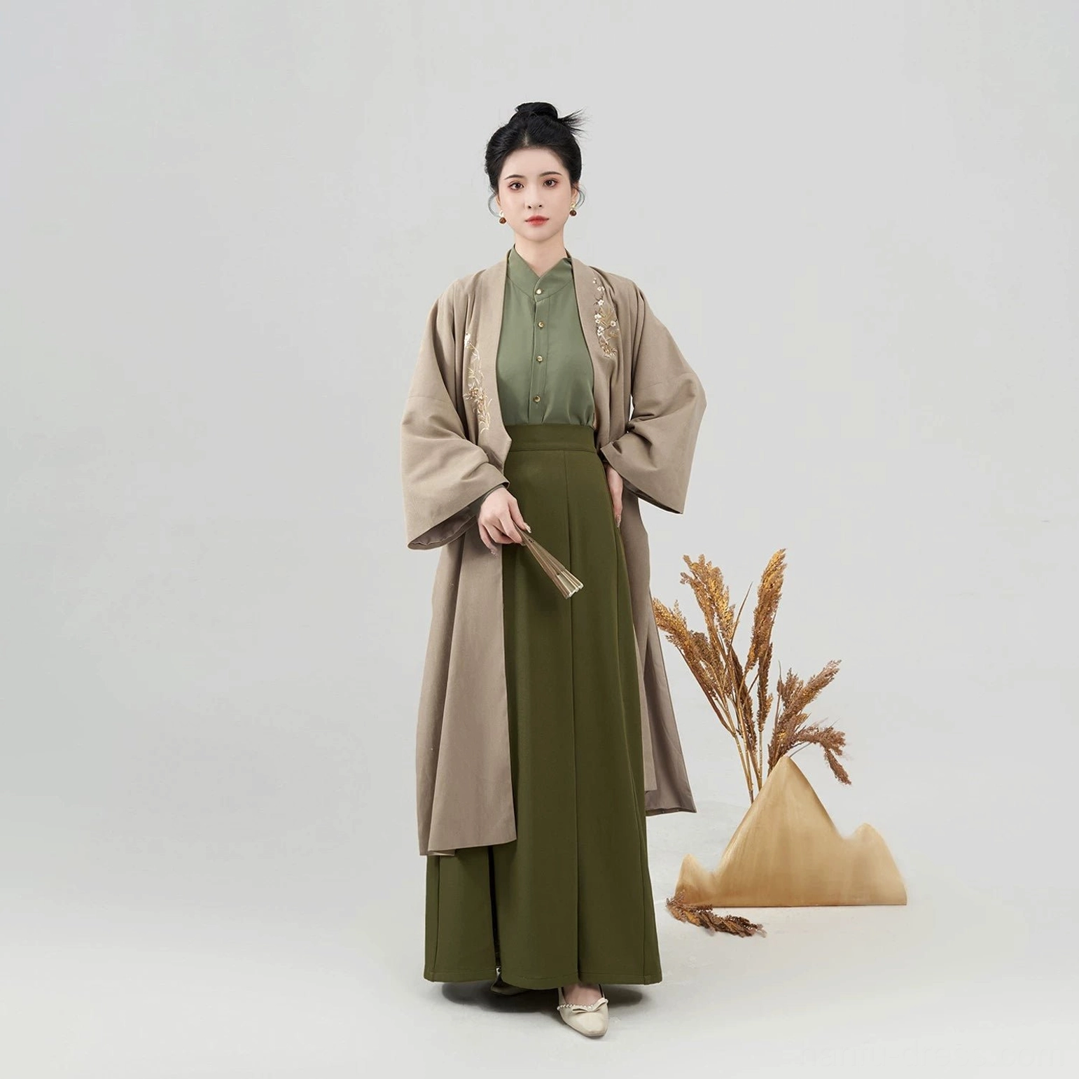 Close front view of a woman wearing a complete modern Song Dynasty Hanfu set with khaki embroidered coat, green top and pleated skirt, holding a folding fan