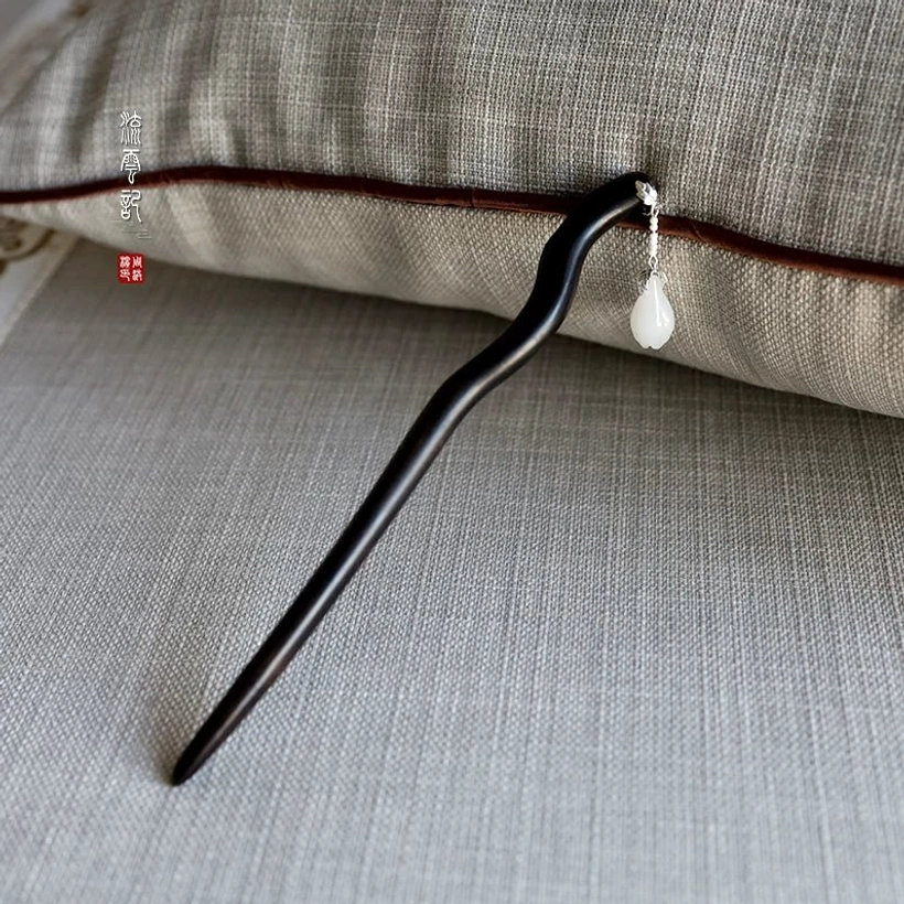 Side view of an ebony Chinese hair stick with a curved black wooden shaft, silver hardware, and teardrop white jade pendant on a linen cushion