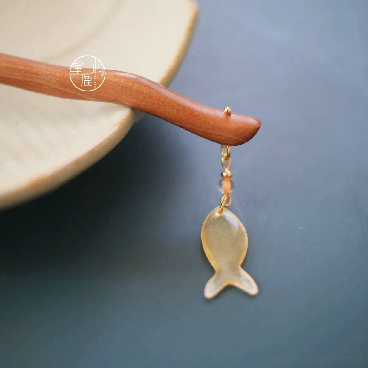 Curved natural wood Chinese hair stick with translucent fish charm and gold-tone chain displayed on a ceramic plate