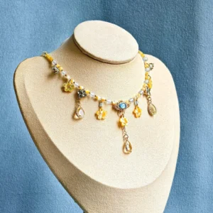 Front three-quarter view of a traditional Hanfu yingluo Chinese necklace with yellow flower charms, faux pearls, blue center gemstone, gold-tone chain, and teardrop pendants on a beige jewelry mannequin