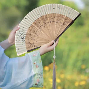 Traditional Chinese folding hand fan with beige paper surface, bamboo ribs, ink-painted reeds and flying birds, carved rib details, and long tassel with jade pendant held with light blue embroidered Hanfu outdoors