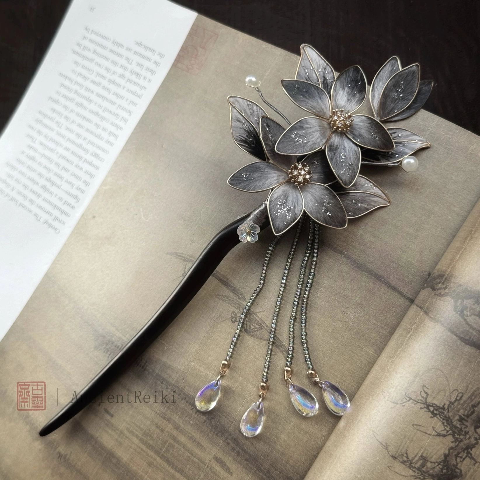 Danqing lotus tassel Chinese hair stick displayed on an open classical Chinese landscape book, showing dark grey flowers with gold edges and iridescent bead pendants