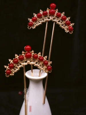 Pair of red and gold Chinese bridal hair pins displayed on a white porcelain vase against a black background