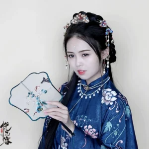 Woman wearing a dark blue Ming-style Hanfu with embroidered lotus details and Chinese tassel hair pin accessories while holding a traditional Chinese hand fan