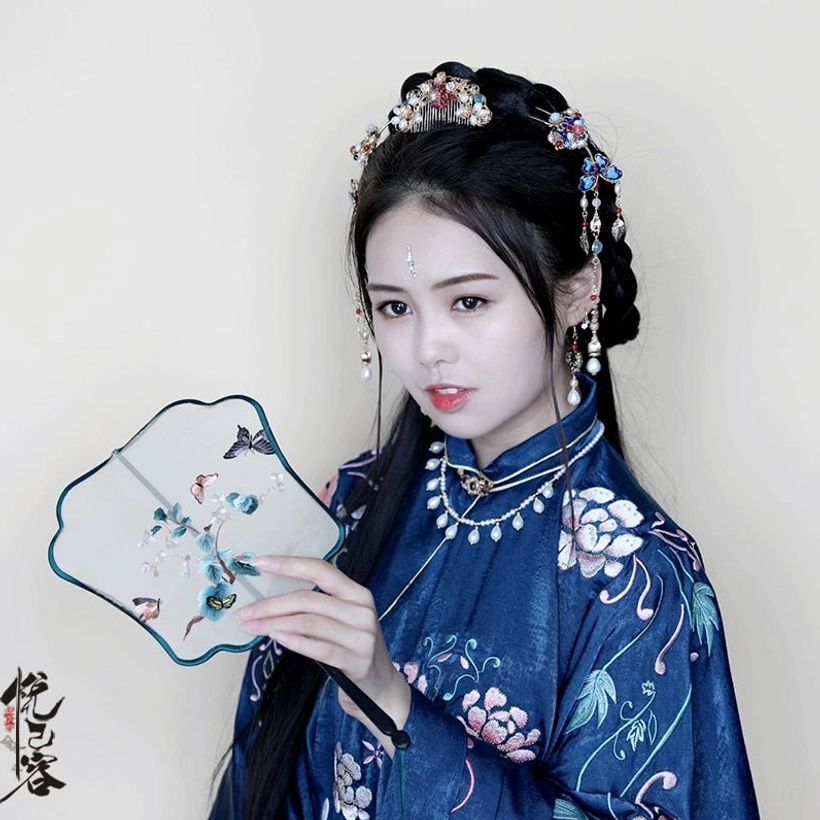 Woman wearing a dark blue Ming-style Hanfu with embroidered lotus details and Chinese tassel hair pin accessories while holding a traditional Chinese hand fan
