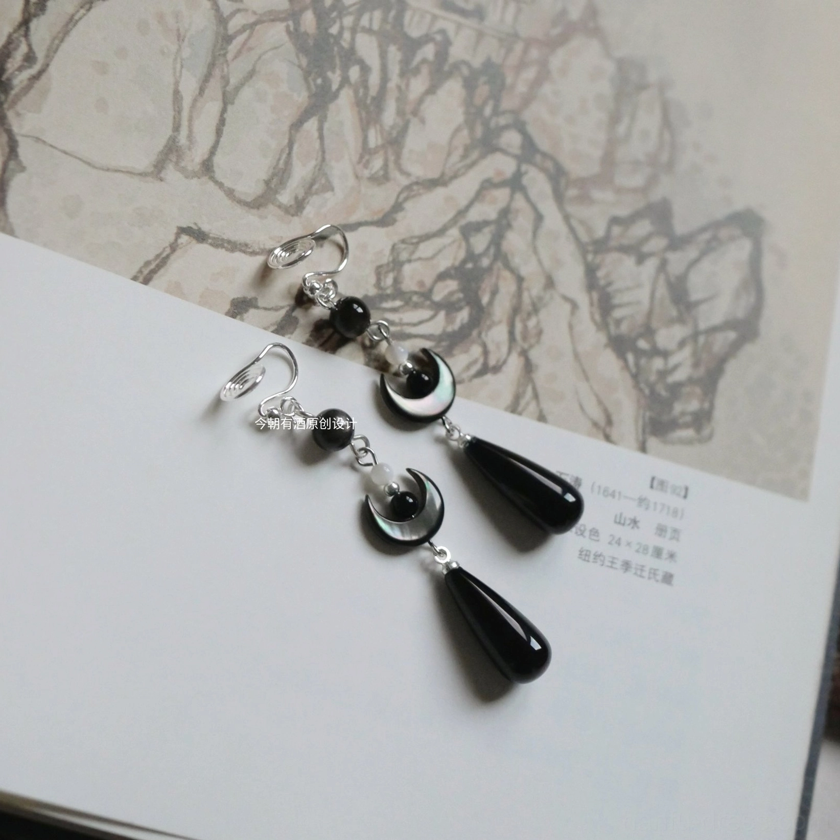 Dark crescent Chinese Hanfu clip-on drop earrings with black teardrop pendants displayed on a traditional Chinese landscape painting page