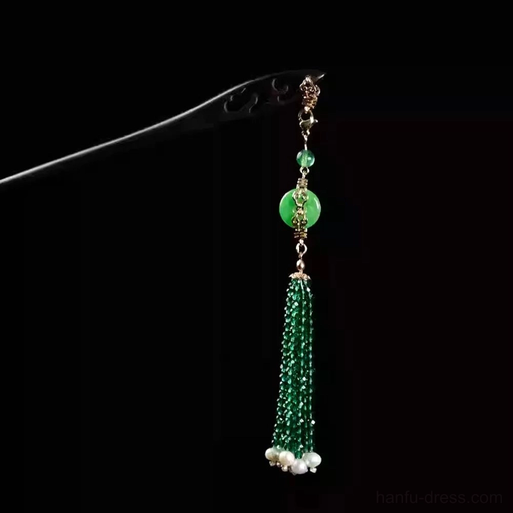 Dark ebony-style Chinese hair stick with emerald green tassel pendant, round peace-buckle charm, white pearl-like decorations, and gold-toned hardware