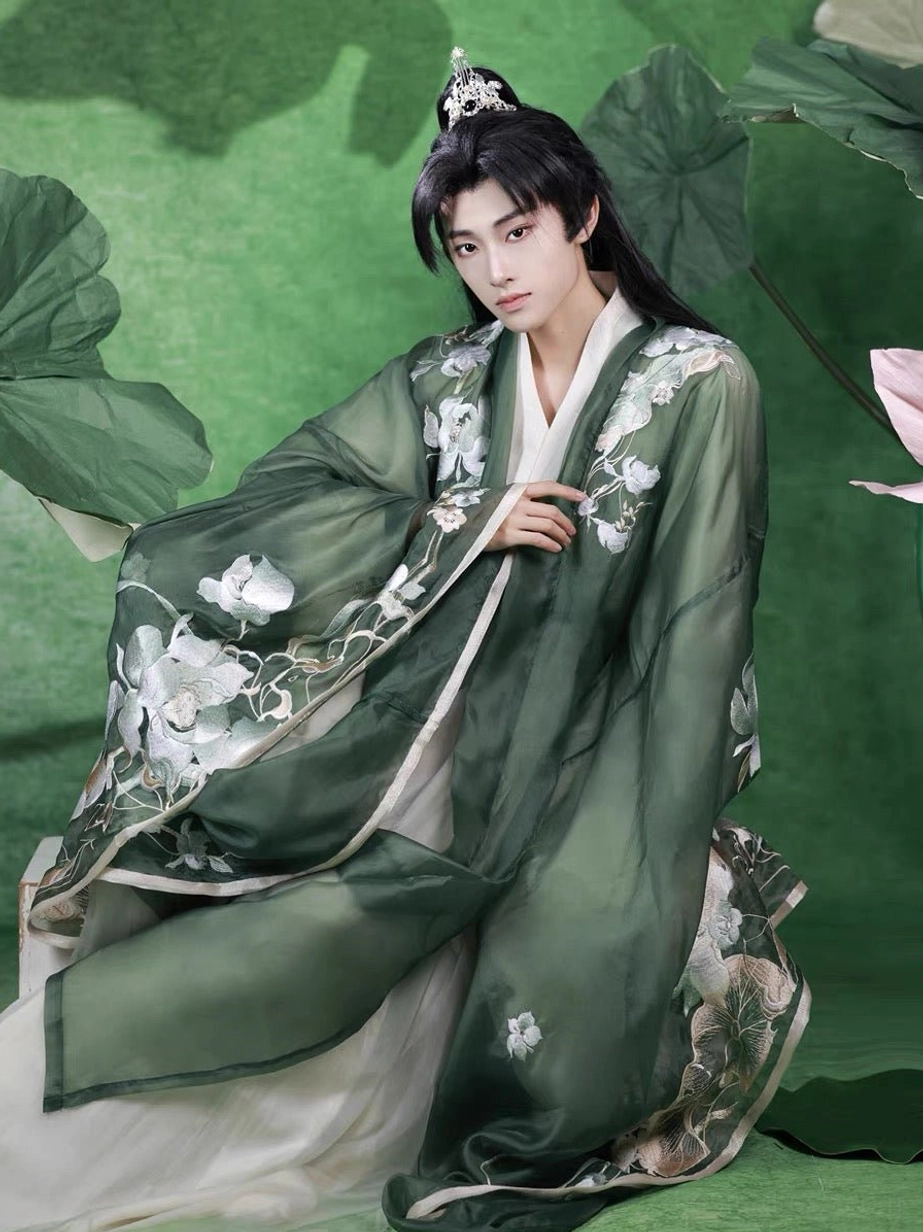 Full-body low-angle view of a male model kneeling in a dark green embroidered Hanfu coat with wide sleeves, large lotus motifs, ivory layered robes, and xianxia styling