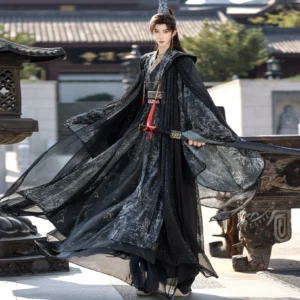Three-quarter front view of a male model wearing a dark immortal unisex hanfu cloak robe with black ink-wash prints, sheer flowing cape, wide sleeves, red tassel waist details, and a traditional sword in an ancient Chinese courtyard