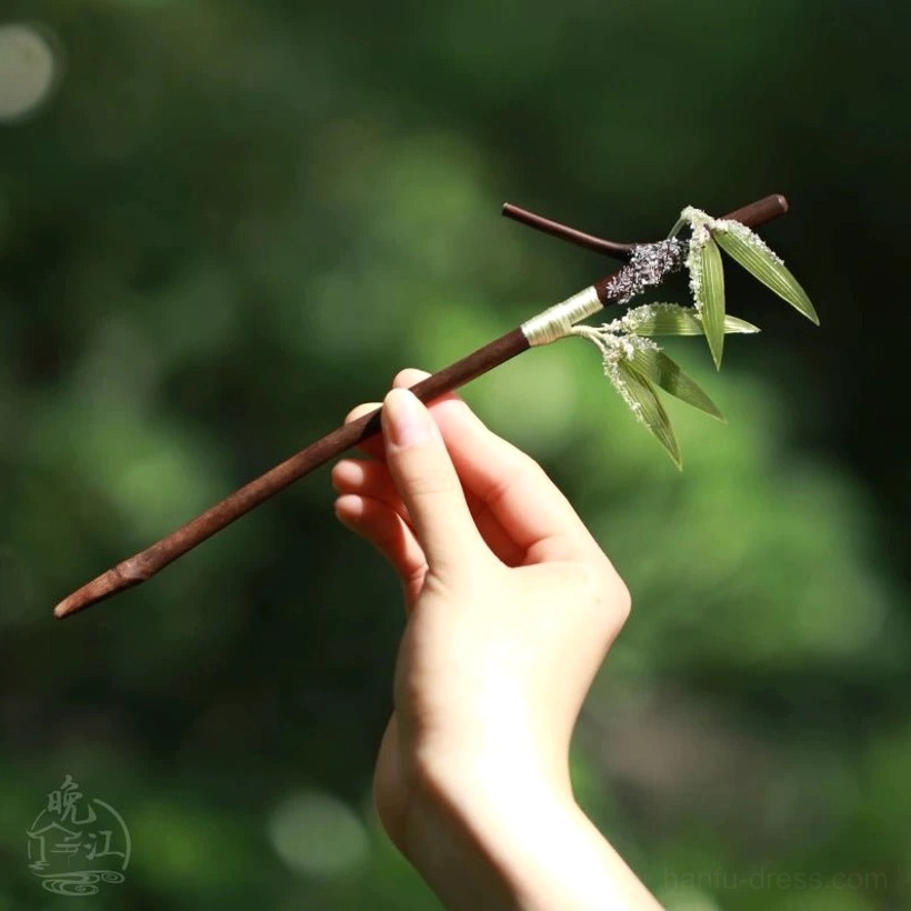 Hand holding a dark wood snow bamboo Chinese hair stick with green leaves faux snow accents and traditional wrapped trim outdoors