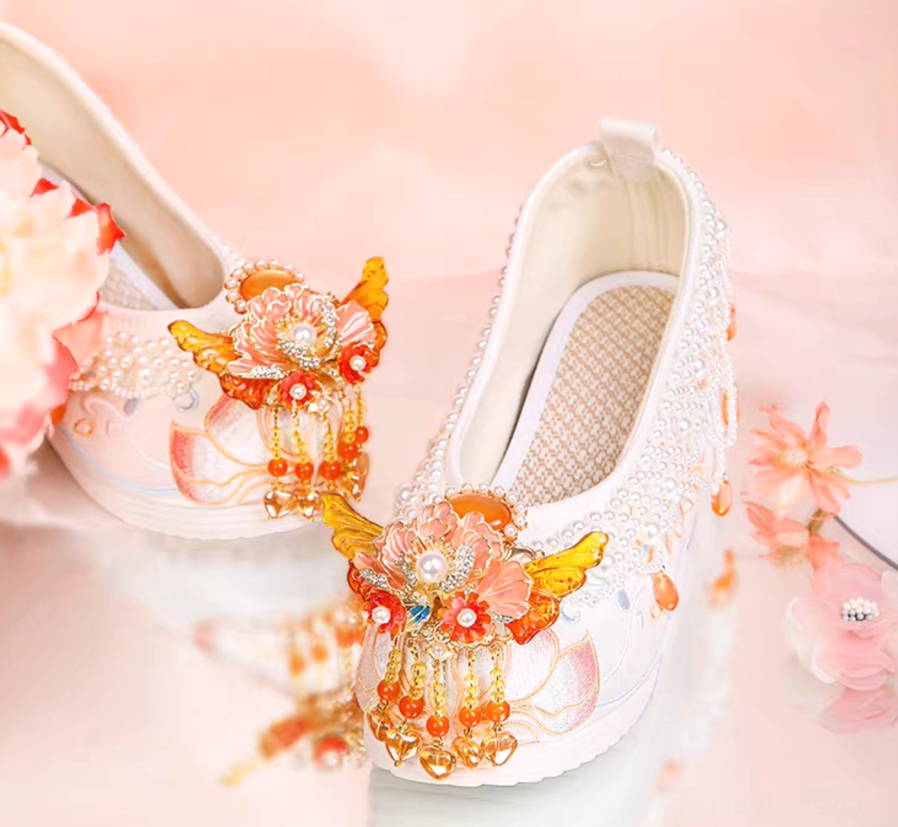 Decorated white Chinese Hanfu shoes with orange-red flower and butterfly tassels, pearl embellishments, and lotus embroidery displayed with floral props