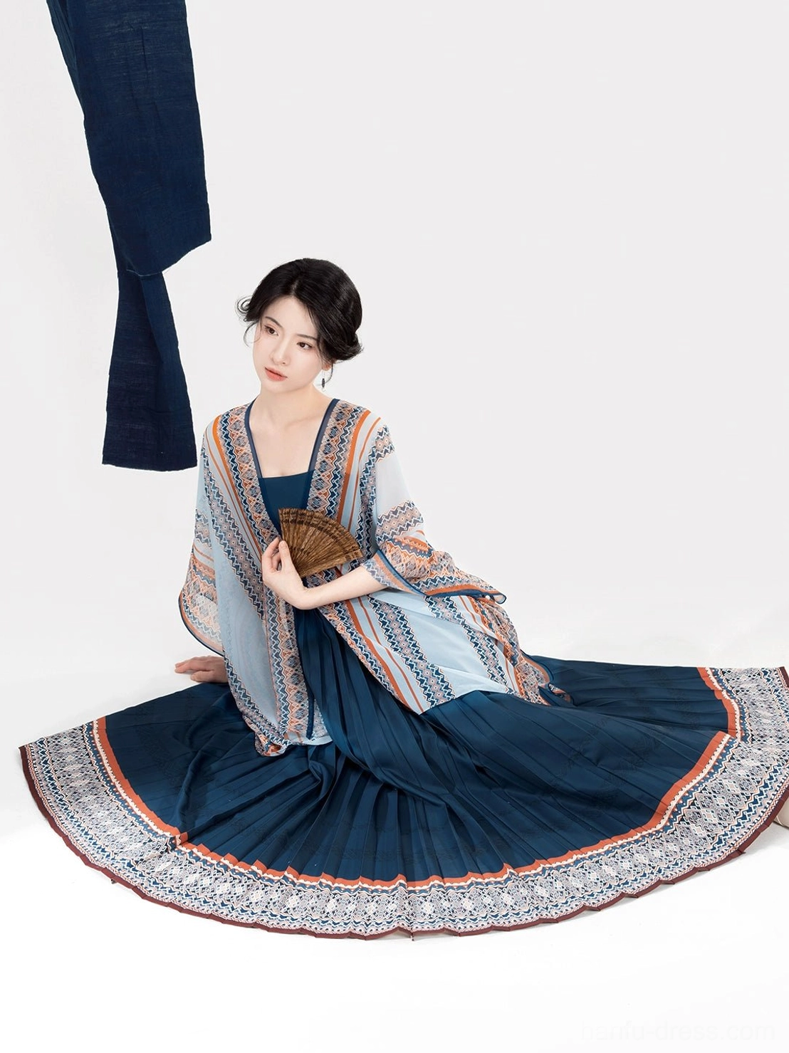 Dong-inspired modern Hanfu two-piece outfit with light blue patterned shawl and dark blue pleated slip dress, shown on a seated woman with traditional updo and wooden fan