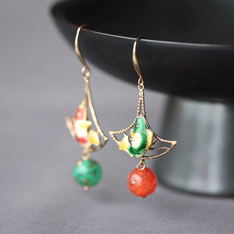 Double Blessing koi earrings displayed on a black bowl with reversed color fish charms, gold-tone frames, and red and green bead pendants