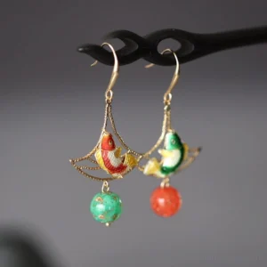 Double Blessing koi fish drop earrings displayed on a black hairpin with gold filigree fan frames, enamel-style koi motifs, green speckled beads, and red bead pendants