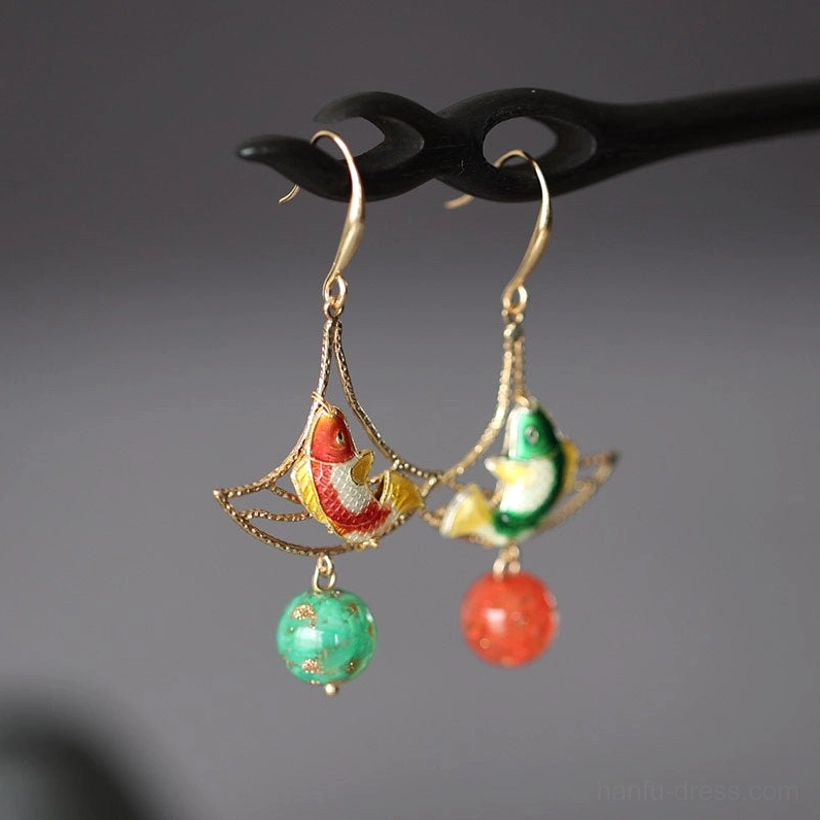 Double Blessing koi fish drop earrings displayed on a black hairpin with gold filigree fan frames, enamel-style koi motifs, green speckled beads, and red bead pendants