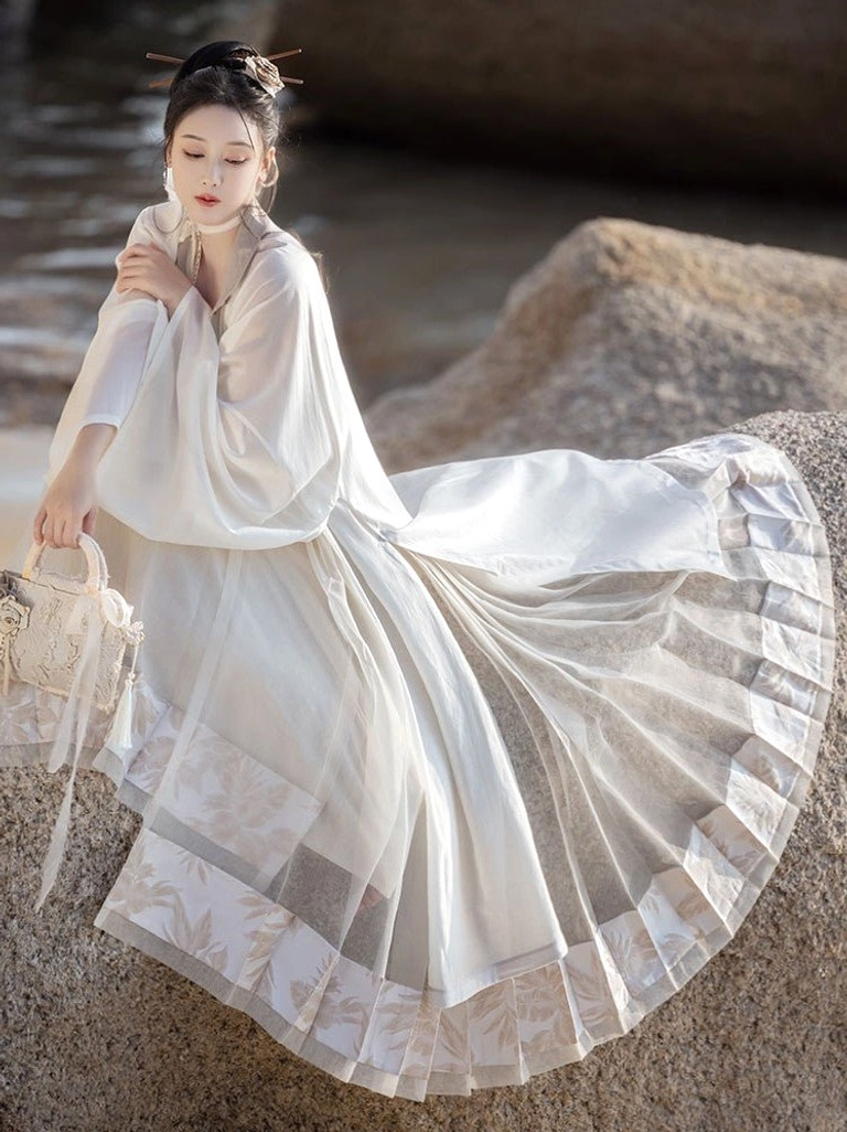 Dream of Mountains Chinese Hanfu set shown in a seated three quarter view beside seaside rocks, featuring sheer flowing layers, embroidered ruffled skirt hem, wide sleeves, and woven handbag styling