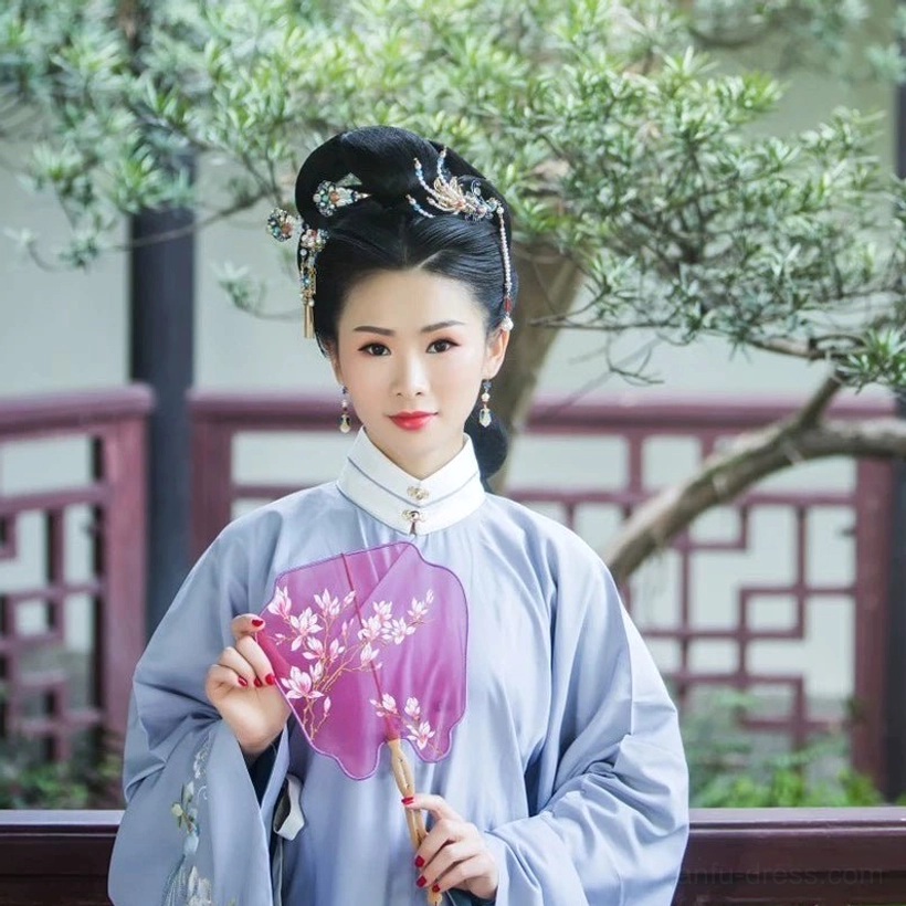 Woman wearing light blue Ming style Hanfu with Dream of Red Mansion phoenix tassel hairpins and purple Chinese folding fan in a traditional garden