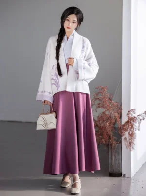 Model wearing a Dreamy Iris Song Dynasty modern hanfu outfit with an ivory embroidered jacket, deep purple pleated wrap skirt, and beige heels in a studio setting