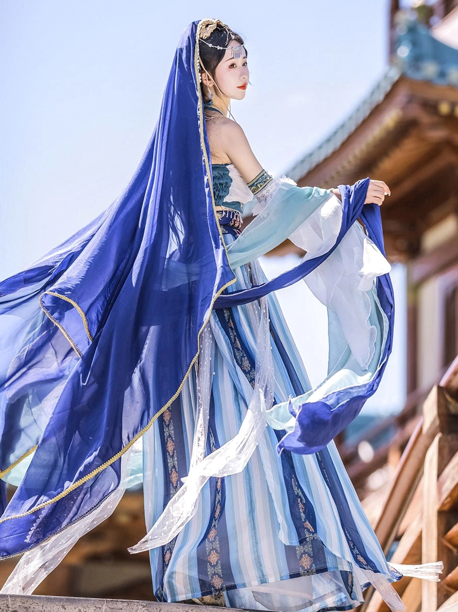 Side back view of royal blue Dunhuang Feitian Hanfu costume showing the long flowing veil, gold-edged garment details, pleated skirt, and trailing sleeve ribbons beside an ancient wooden pavilion