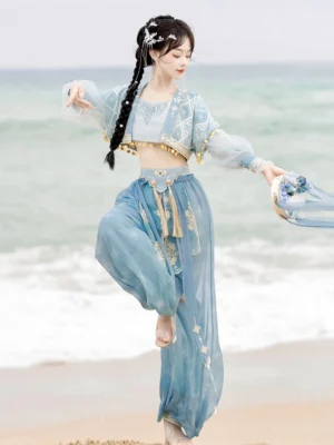 Sky blue Dunhuang Feitian Tang Dynasty Hanfu dance costume with gold trim, sheer lantern sleeves, cropped top, and flowing silhouette worn by a model at the seaside