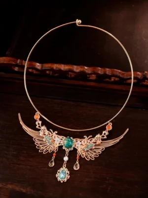 Top-down flat lay of a Dunhuang-inspired Chinese necklace on a dark polished wooden surface, showing the open gold-tone collar ring, matching winged pendant, decorative beads, and hanging ornaments