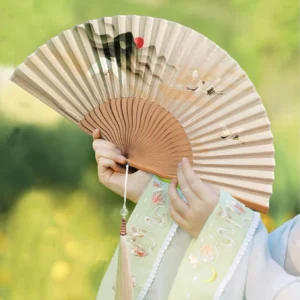 Dusk crane landscape Chinese folding hand fan with red sun print, wood-toned bamboo ribs and beaded tassel held with embroidered Hanfu sleeves outdoors
