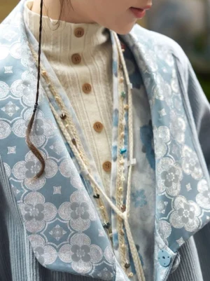 Close-up detail of the dusty blue corduroy coat collar showing silver blue floral jacquard shawl trim, soft ribbed fabric texture, round button fastening, and layered necklace styling