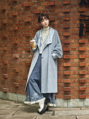 Full body view of a woman wearing a dusty blue corduroy Hanfu-inspired Chinese style trench coat with a silver floral jacquard shawl collar, matching cuffs, round fabric buttons, and midi length silhouette outdoors