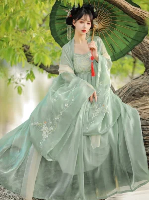 Three-quarter front view of a glossy dusty green Tang Dynasty style Hanfu dress with floral embroidery, flowing sleeves, traditional hairstyle, and red tassel oil-paper umbrella near the water