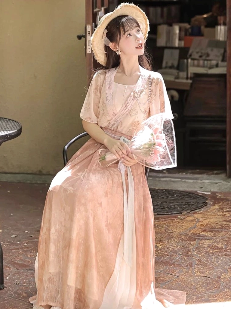 Seated view of a dusty pink gradient floral modern Hanfu dress with flowing maxi skirt straw hat and romantic outdoor cafe styling