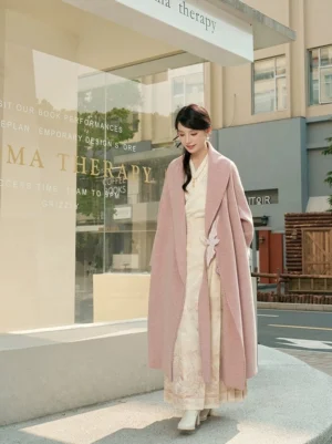 Dusty pink women's Hanfu-inspired double-sided winter coat with shawl lapel, waist sash, crane embroidery buckle, and long flowing silhouette styled over a cream Hanfu dress