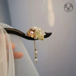 Close-up of a traditional ebony Chinese hair stick with curved wooden shaft, floral ornament, green beads, pearl accents, and long hanging tassel displayed against sheer fabric