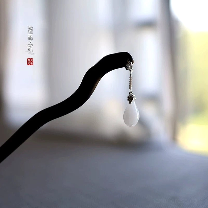 Close-up of a handmade ebony Chinese hair stick with a wavy black wooden shaft, silver-toned chain, and white magnolia-shaped jade pendant on soft fabric