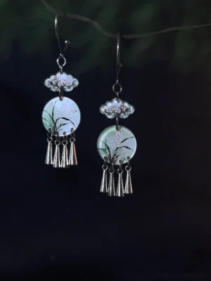 Elegant Chinese earrings with ink-painted orchid pendants, silver-tone cloud details, and hanging tassels displayed against blurred dark green foliage