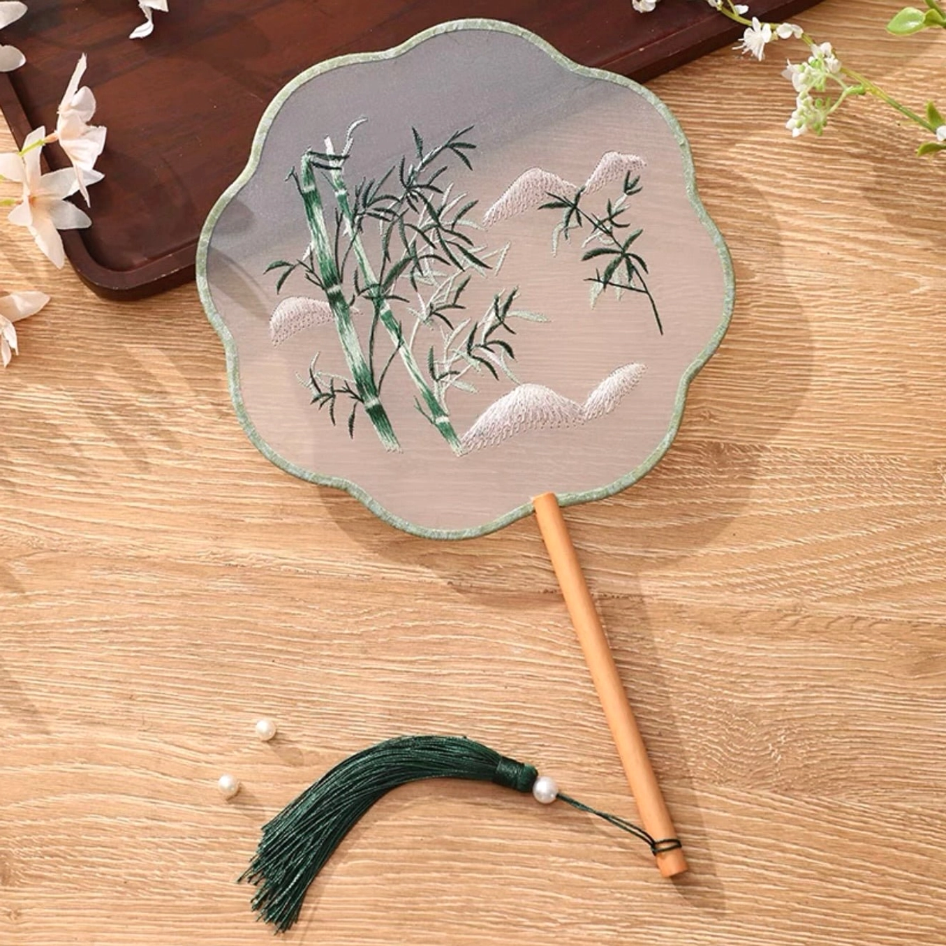 Embroidered bamboo and snow Chinese round hand fan tuanshan with scalloped light green trim, sheer fabric surface, bamboo handle, dark green tassel, and pearl pendant displayed on a wooden table