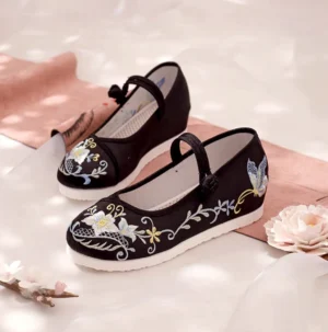 Embroidered black Chinese Hanfu cloth shoes with floral and butterfly embroidery, buckle ankle straps, and white wedge rubber soles displayed in a traditional Chinese style setting