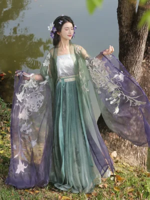 Front three-quarter view of an embroidered Chinese hanfu outfit with sheer floral cardigan, white waist belt, camisole, and green purple gradient wrap skirt beside a tree