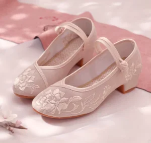 Pair of light apricot pink embroidered Chinese Hanfu shoes with floral detailing, Chinese knot ankle straps, and low block heels displayed on a soft pink traditional-style backdrop