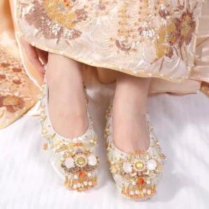 Close-up of embroidered off-white Chinese Hanfu wedding shoes with champagne floral beadwork, warm gemstone accents, metallic ornaments, and dangling tassels worn with a traditional Xiuhe dress