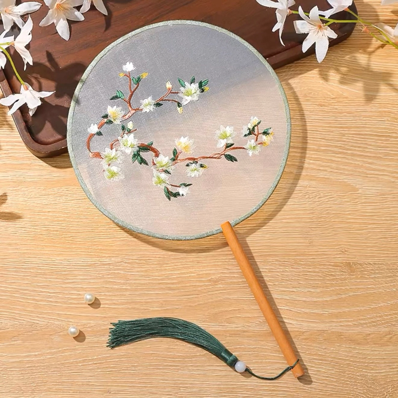 Embroidered Chinese round hand fan with white flowering branch design, light green fabric edging, wooden handle, dark green tassel, and white bead ornament displayed on a wooden table