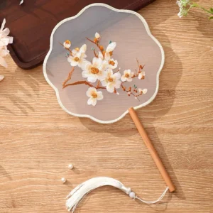 Embroidered Chinese tuanshan round hand fan with translucent floral surface, scalloped edge, wooden handle, and white tassel displayed on a warm wooden table