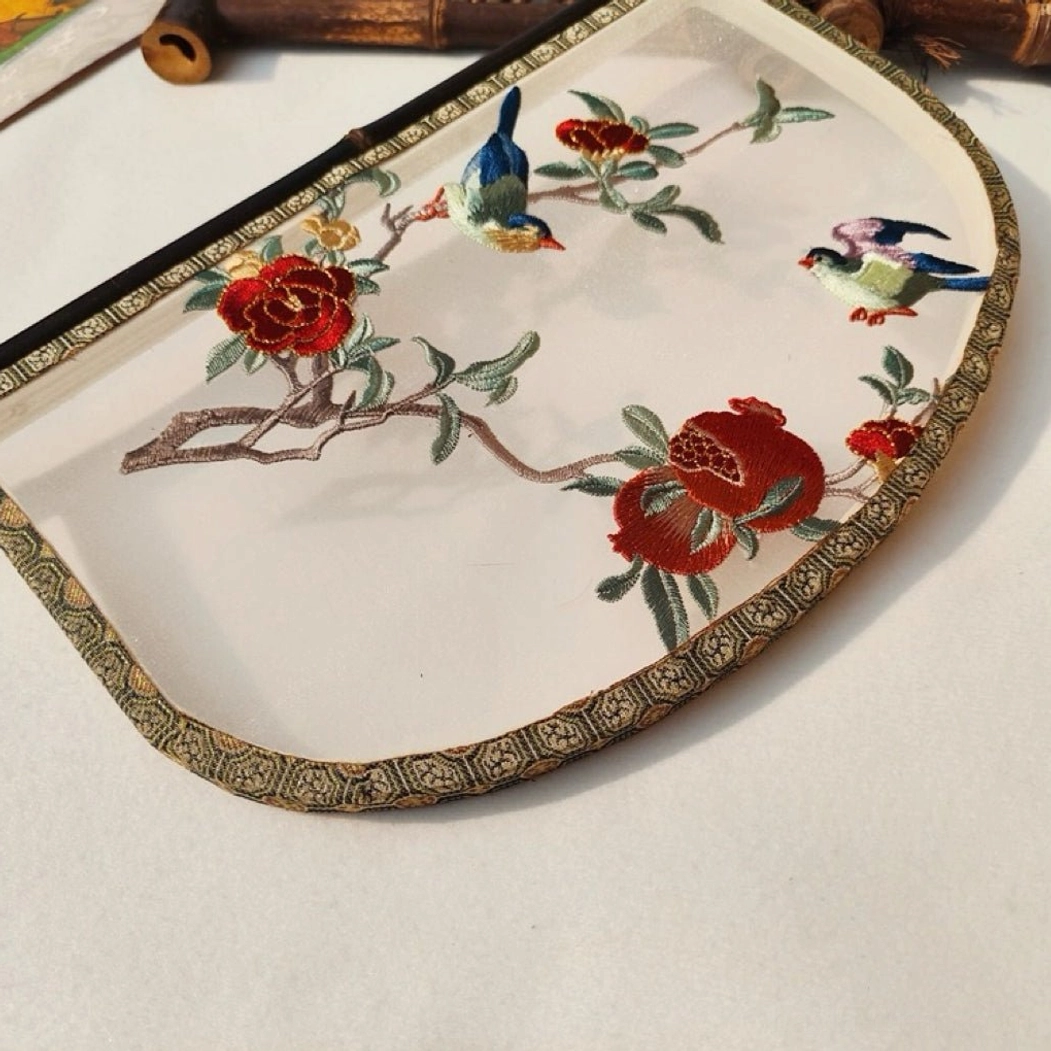 Close-up front view of a decorative knife-shaped Chinese hand fan showing colorful embroidered songbirds, red flowers, green branches, pomegranates, translucent fabric, and geometric brocade-style trim