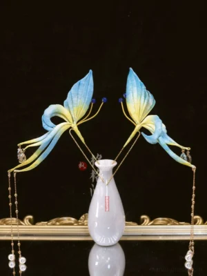 Pair of emerald butterfly Hanfu hair pins with gradient blue and golden wings, blue beads, gold chains, pearl pendants, and crystal tassels displayed on a porcelain vase and gold tray