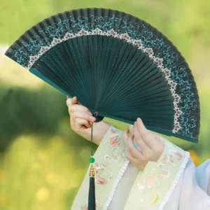 Emerald green Chinese folding hand fan with blue vine pattern white lace trim and green tassel held with embroidered Hanfu outdoors