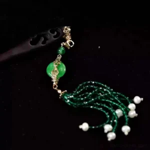 Close-up of an emerald green Chinese hair stick with jade-style peace-buckle pendant, pearl-like accents, gold-toned connectors, and dark tassel on a black background