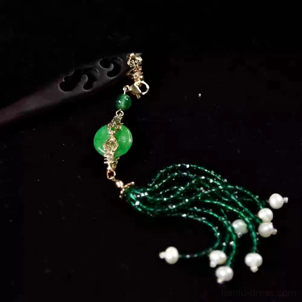Close-up of an emerald green Chinese hair stick with jade-style peace-buckle pendant, pearl-like accents, gold-toned connectors, and dark tassel on a black background