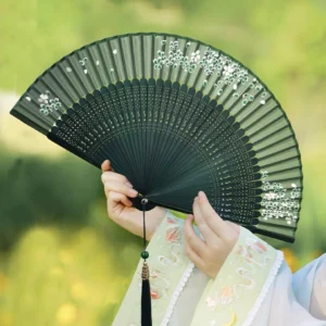 Emerald green traditional Chinese folding hand fan with white cherry blossom embroidery, carved dark bamboo ribs, black tassel, and jade ornament held with Hanfu styling outdoors