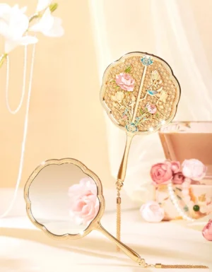 Empress Garden gold toned filigree Chinese hand mirror displayed on a warm vanity table with pink flowers, scalloped frame, enamel floral back, and hanging tassel