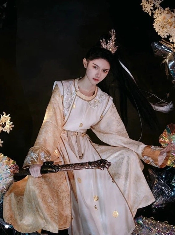 Eternal Dawn Tang Dynasty hanfu robe in light gold and beige with dragon-inspired embroidery, round neckline, metal waist belt, traditional hair crown, and antique sword