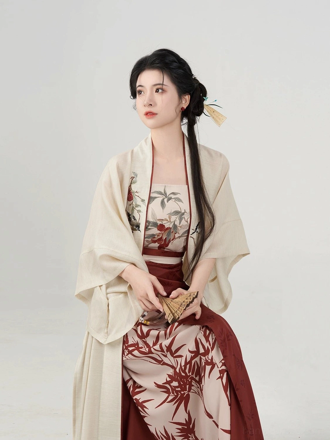Evening Magpie Song-style Hanfu seated side view with a beige outer robe, bamboo print skirt, and traditional folding fan